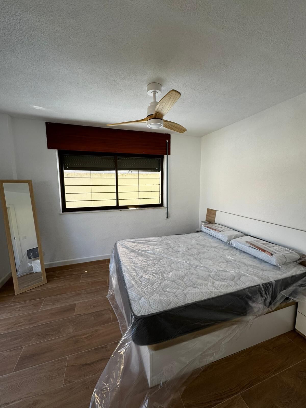 A bedroom with a bed, a ceiling fan, a window with blinds, and a wooden floor.