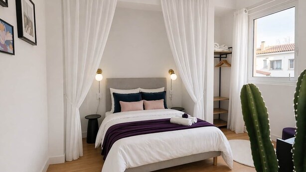 Un dormitorio con cama, cortinas blancas, una mesita de noche, un cactus y una ventana con vistas.