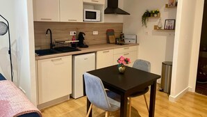 Una cocina compacta con gabinetes blancos, un microondas, una cafetera y un lavamanos. Hay una pequeña mesa de comedor con dos sillas y un florero con flores.