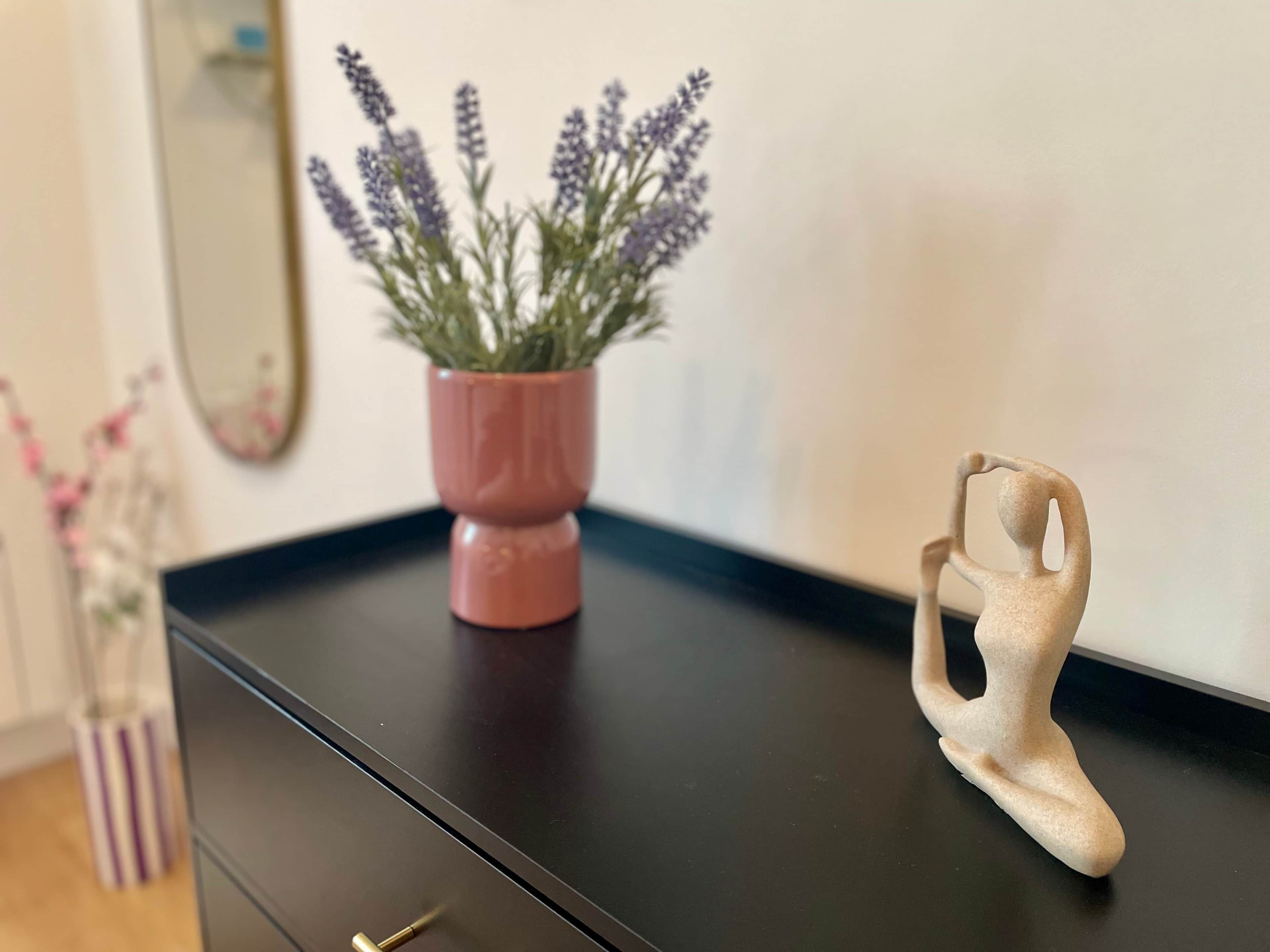 Une commode noire ornée d’un vase rose contenant des fleurs de lavande et d’une petite sculpture abstraite.