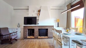 Een woonkamer met een open haard, een televisie, een eettafel met stoelen en een raam met gordijnen.