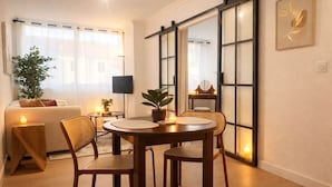 Un salon chaleureux avec une table ronde en bois, deux fauteuils en osier, un canapé, un téléviseur et une grande fenêtre ornée de rideaux légers.