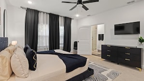 Un dormitorio con cama, ventilador de techo, televisor, cómoda y puerta que da a un baño.