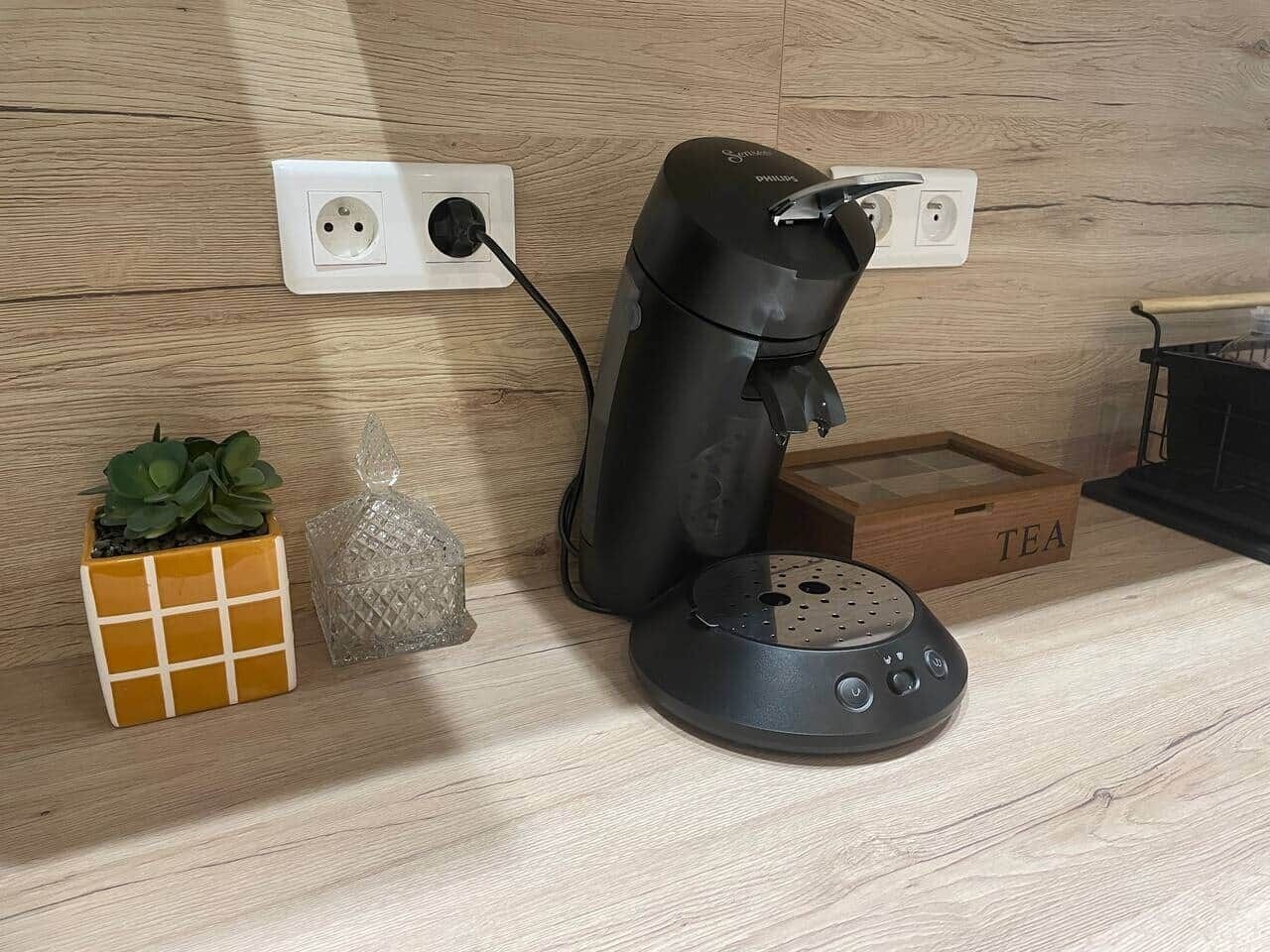 Una cafetera sobre una encimera de madera, una pequeña planta en maceta, un recipiente de vidrio y una caja de madera con la etiqueta 'TEA'.