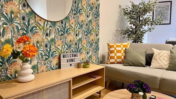 Un salon avec un papier peint à motifs floraux, un miroir rond, une pancarte « BIENVENUE À LA MAISON », un canapé avec des coussins à carreaux, une table basse avec un vase et une télécommande, et une bibliothèque.