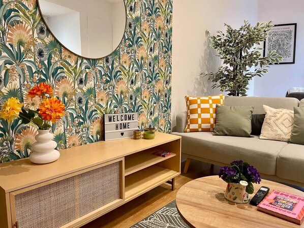 Una sala de estar con papel tapiz floral, un espejo redondo, un cartel que dice 'WELCOME HOME', un sofá con cojines a cuadros, una mesa de centro con un jarrón y un control remoto, y una estantería.
