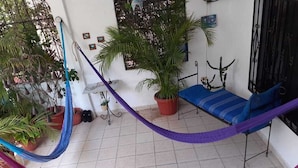 Una hamaca en una habitación con suelo de baldosas, una cama con colcha a rayas azules y blancas, y varias plantas en macetas.