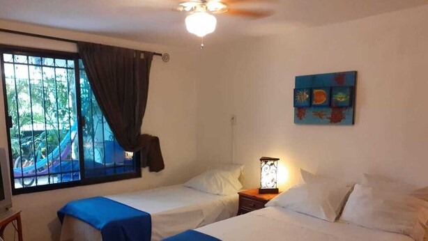 Una habitación con dos camas, una ventana con cortinas, un ventilador de techo, una lámpara fijada a la pared y un cuadro de colores.