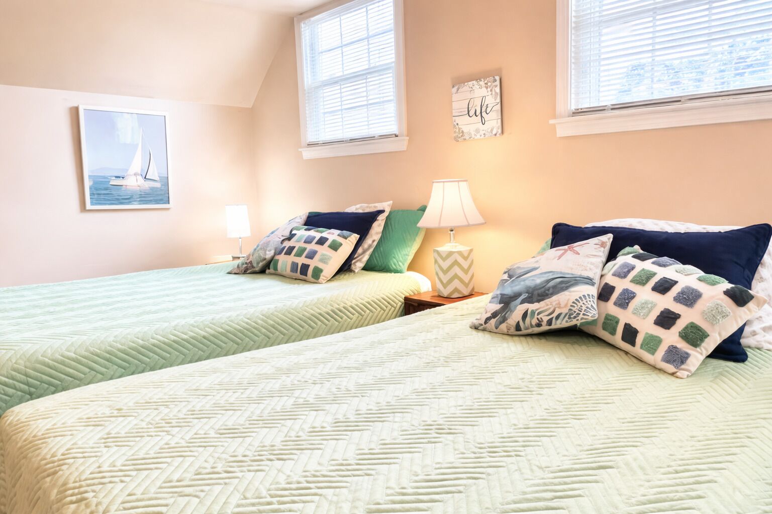 A bedroom with two beds, each with patterned bedding and pillows, a bedside table with a lamp, and a framed picture on the wall.