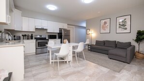 A modern kitchen with white cabinetry, a dining area with a white table and chairs, a grey sofa, and two framed pictures on the wall. - Modern Comfort 2BR in Delta - Near Parks & Shops (Delta)