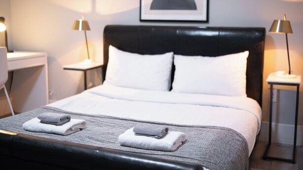 A neatly made bed with white linens and two folded towels, a black headboard, and two bedside tables with lamps. - Modern Comfort 2BR in Delta - Near Parks & Shops (Delta)