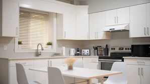 A modern kitchen with white cabinetry, a dining table, chairs, and a window with blinds. - Modern Comfort 2BR in Delta - Near Parks & Shops (Delta)