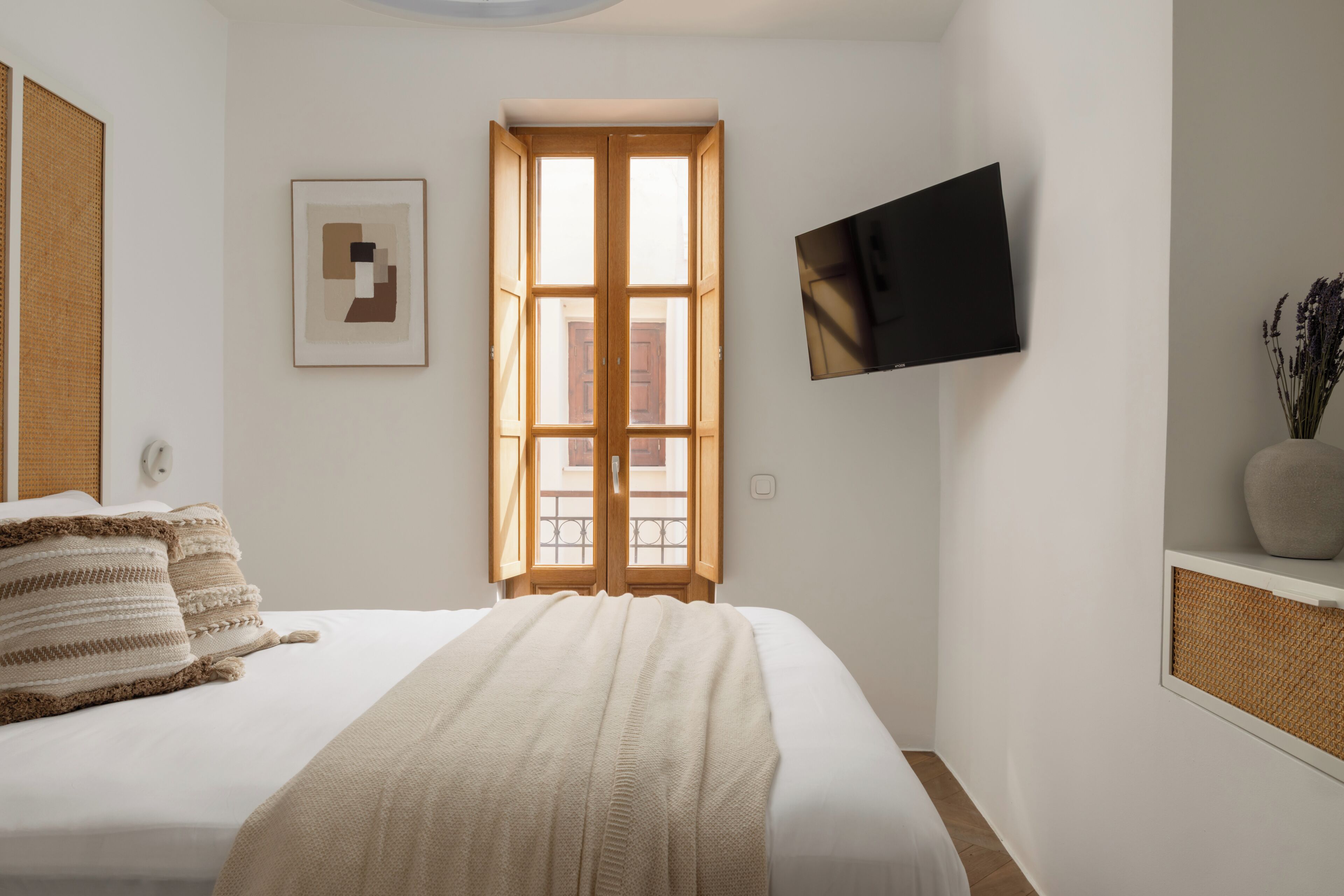 A bedroom with a bed, a window, a flat-screen TV, and a framed picture on the wall.