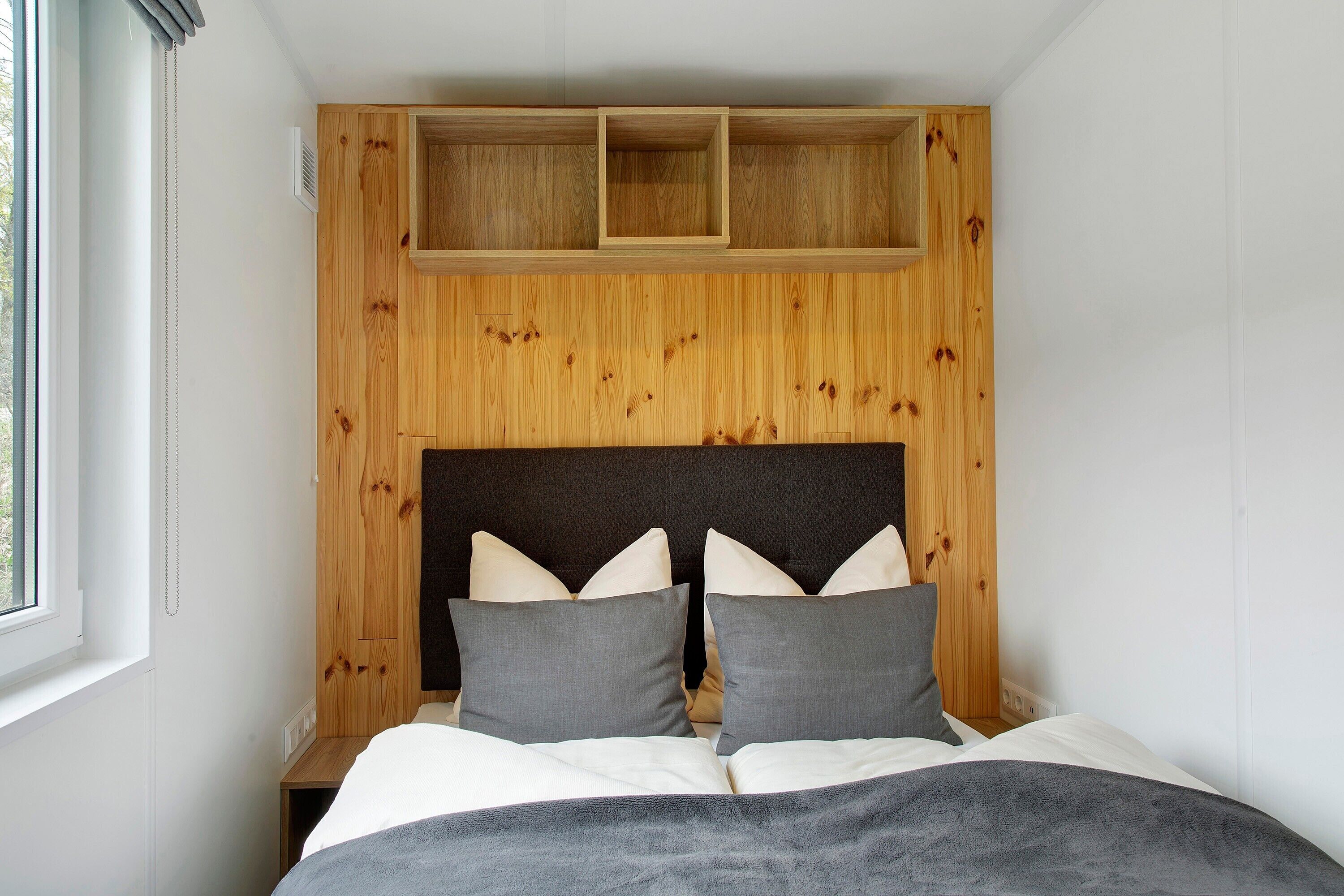 A bedroom with a wooden headboard, a bed with grey bedding, and a window on the left.