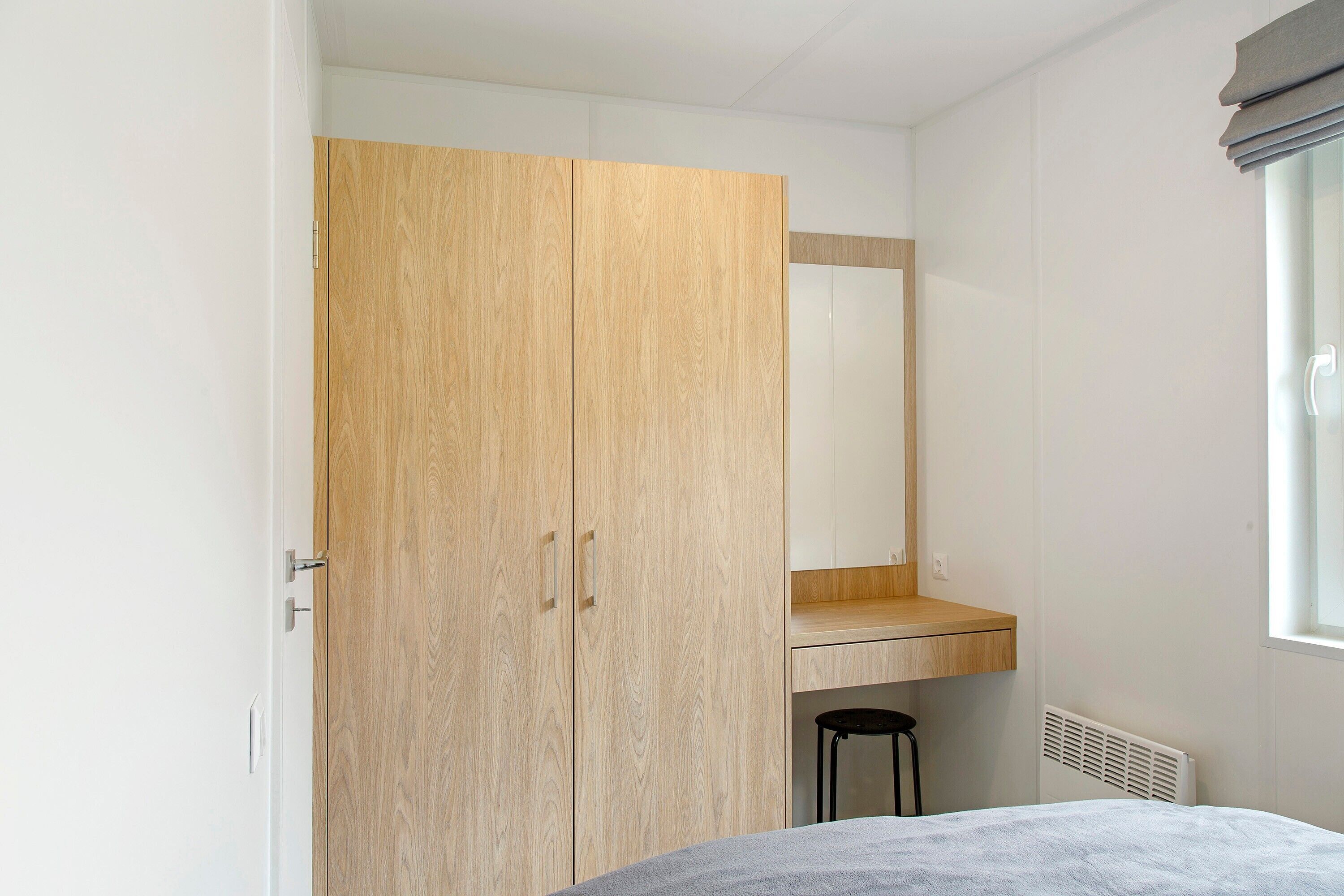 A bedroom with a wooden wardrobe, a small stool, a bed, and a window.