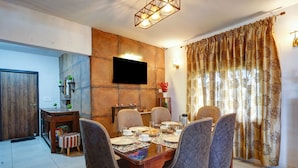 A dining area with a wooden table set for four, surrounded by cushioned chairs, a flat-screen TV mounted on a stone wall, and a patterned curtain on the window. - Panoramic Views 4-bedroom Luxury villa in marvelous Mukteshwar for your getaway (Mukteshwar)