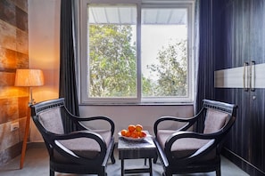 Une pièce avec deux chaises, une table sur laquelle se trouvent des fruits, une lampe et une fenêtre donnant sur des arbres.