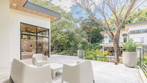 A modern outdoor patio with white furniture, a glass wall, and a view of a garden and neighboring buildings.