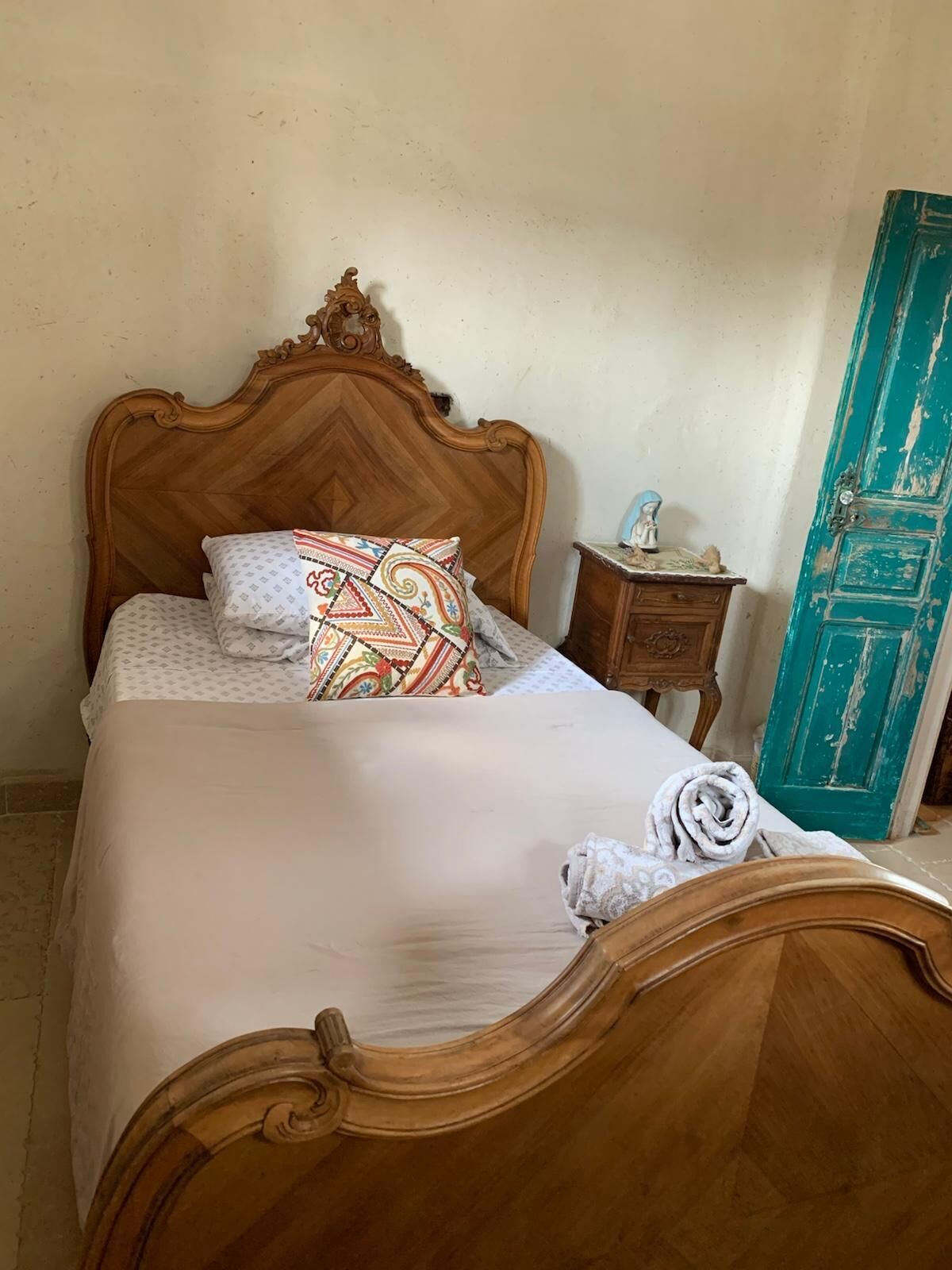 A bed with a wooden headboard, a patterned pillow, a white blanket, and a rolled towel.