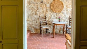 Una puerta abierta que da a un cuarto de paredes de piedra, con una mesa redonda de fibras naturales, dos sillas y una pequeña planta en maceta.