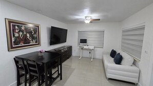 Ein Wohnzimmer mit Fernseher, einem Esstisch mit Stühlen, einem weißen Sofa und einem Deckenventilator.
