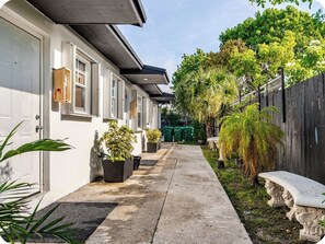 A narrow pathway leading to a row of white buildings with windows, surrounded by greenery and a wooden fence. - Miami Health District 1/1 w/2Gbps WiFi Stainless Steel 75” & 55” TV Pet friendly (Miami)
