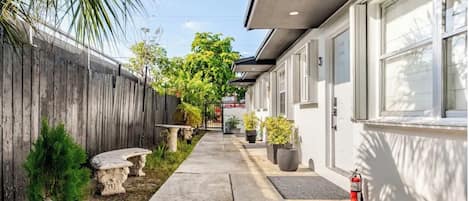 A narrow pathway leading to a building with a white exterior, a wooden fence on one side, and potted plants along the path.