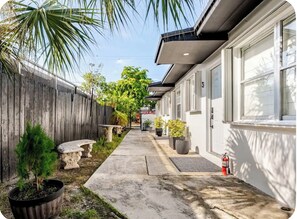 A narrow pathway leading to a building with a white exterior, a wooden fence on one side, and potted plants along the path. - Miami Health District 1/1 w/2Gbps WiFi Stainless Steel 75” & 55” TV Pet friendly (Miami)