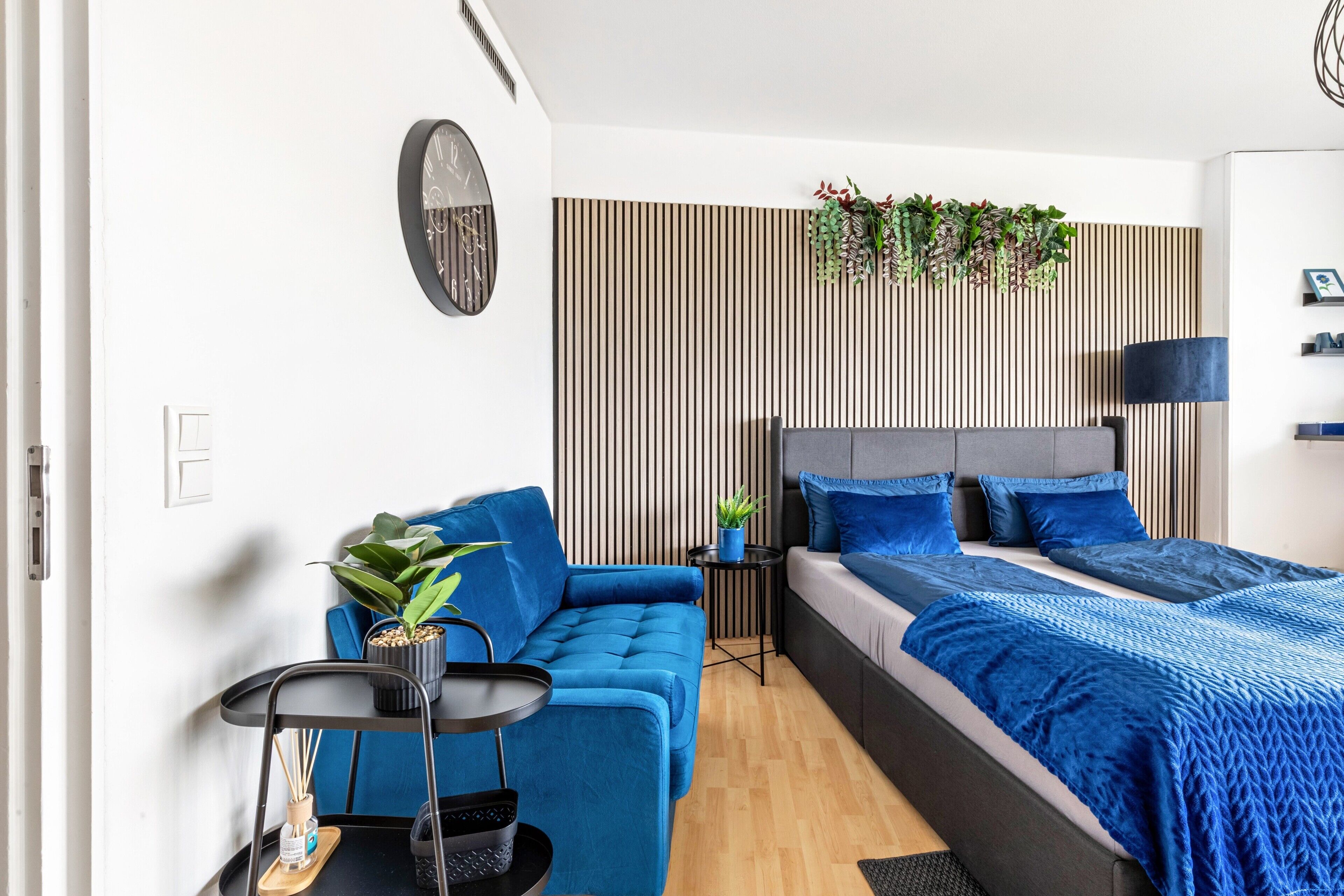 Une chambre moderne avec un canapé bleu, un lit avec une literie bleue, une petite table d’appoint et une horloge murale.