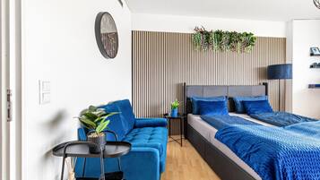 Une chambre moderne avec un canapé bleu, un lit avec une literie bleue, une petite table d’appoint et une horloge murale.