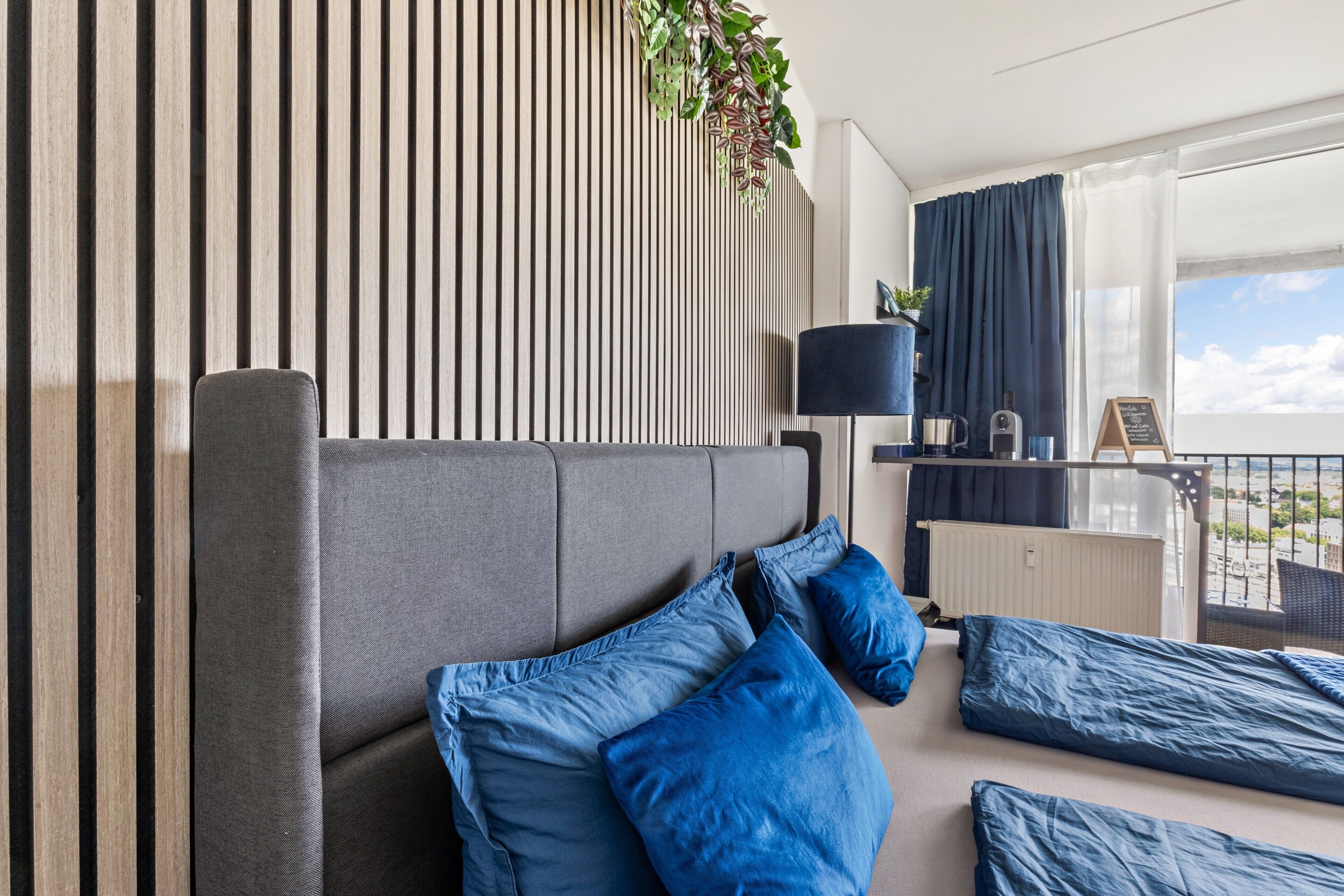 Ein modernes Hotelzimmer mit grauem Sofa, blauen Kissen, einem kleinen Tisch mit Lampe und Blick auf den Himmel vom Balkon aus.