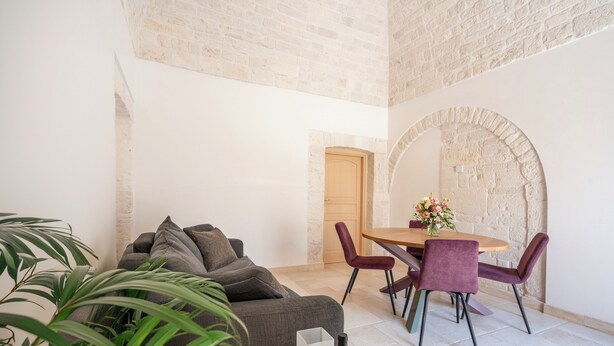 A room with a brick wall, a wooden door, a grey sofa, a dining table with purple chairs, and a potted plant. - Trullo in Alberobello apartment for 4/6 people (Alberobello)