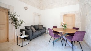 A modern living room with a grey sofa, a round wooden dining table, and purple chairs. There is a potted plant and a small table with a vase of flowers. - Trullo in Alberobello apartment for 4/6 people (Alberobello)