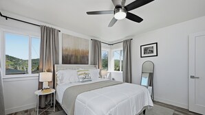 A bedroom with a bed, a ceiling fan, a window with curtains, a bedside table with a lamp, and a framed picture on the wall.