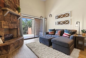 Una sala de estar moderna con chimenea de piedra, un sofá gris con almohadas con la bandera estadounidense y vista a una piscina a través de puertas de cristal.