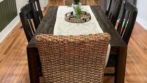 Un coin repas avec une table en bois et des chaises en osier, une cuisine à l’arrière-plan et un réfrigérateur.