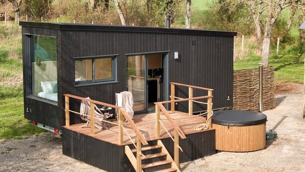 A modern, compact cabin with a deck, a hot tub, and a small outdoor seating area.