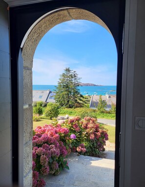 Un arco que da a una vista de la costa, con cielo despejado, un pino y flores rosadas.