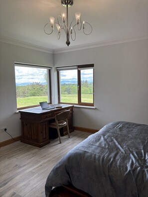 Un dormitorio con una cama grande, un escritorio con una computadora portátil y dos ventanas que ofrecen vista a un paisaje verde.