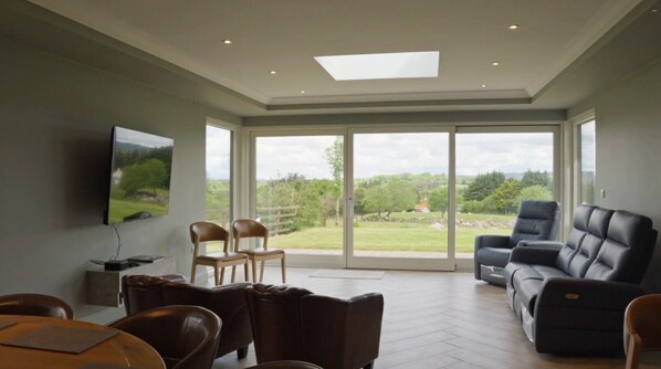 A spacious living room with a large sliding glass door leading to a balcony, a flat-screen TV mounted on the wall, and a comfortable seating area with leather sofas and chairs.