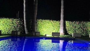 Ein beleuchteter Pool bei Nacht, im Hintergrund zwei Palmen und ein Heckenstreifen.
