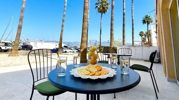 Ein runder Tisch mit Stühlen, einer Fruchtschale und einem Glas Wasser auf einer Terrasse mit Palmen und blauem Himmel.