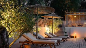 Una terraza de madera con sillones de descanso y un paraguas de paja, rodeada de follaje.