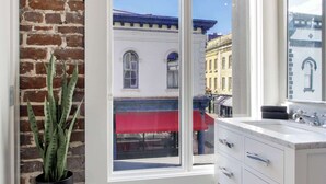 Un bagno con una grande finestra, un mobile lavabo bianco, una pianta in vaso e vista sugli edifici esterni.