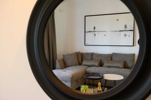 A living room with a sectional sofa, a small round table with a bottle and glasses, and a framed abstract painting on the wall.
