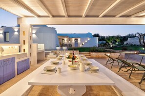 Un espace de restauration en plein air avec une longue table dressée pour un repas, entourée de chaises et d’un coin salon abrité.