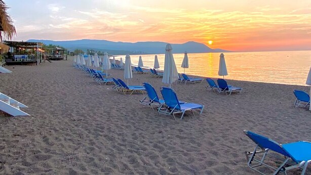A beach with rows of blue lounge chairs and white umbrellas, a thatched-roof structure, and a sunset over the water.