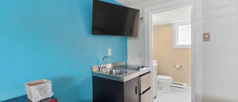 A compact kitchenette with a microwave, sink, and refrigerator, a flat-screen TV mounted above the counter, and a door leading to a bathroom.