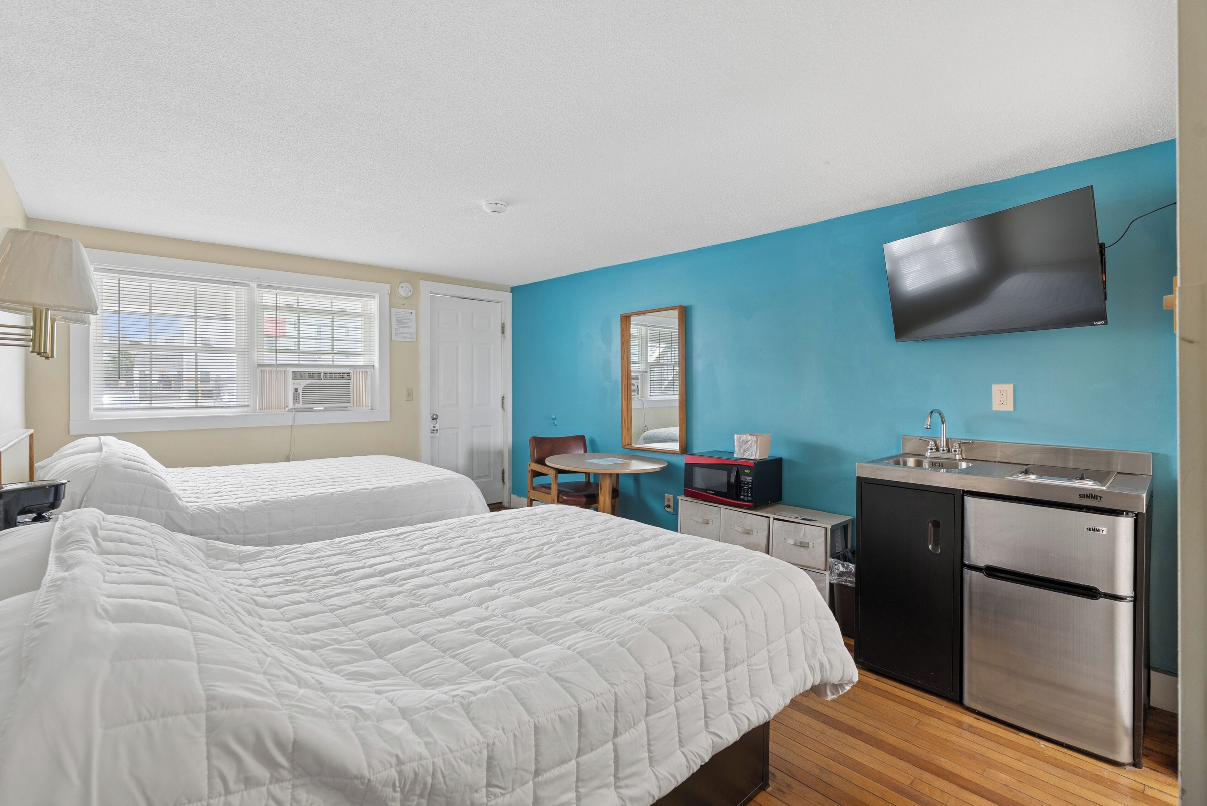 A room with two beds, a kitchenette, a TV mounted on the wall, and a window with blinds.