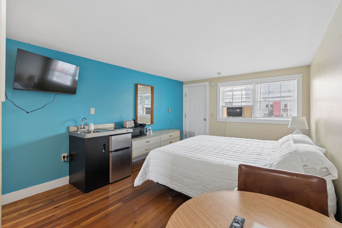 Une chambre d’hôtel équipée d’un lit, d’un bureau, d’une kitchenette, d’une télévision et d’une fenêtre avec des stores.